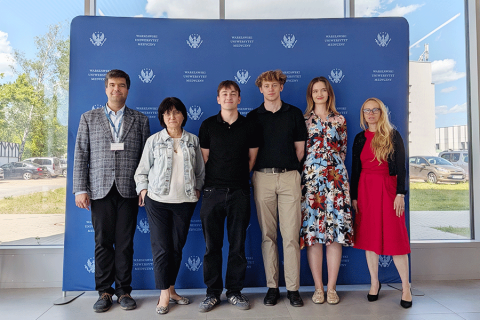 Six people stand side by side in front of a blue backdrop with the Warsaw Medical University logo. The participants are arranged in a row and pose for a photo. Large windows and parts of the outdoor surroundings are visible in the background.