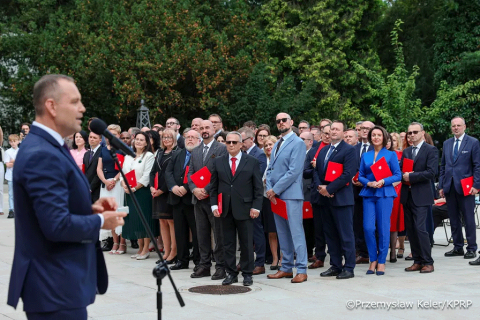 Grupa osób w czasie wręczenia nominacji. Po lewej na pierwszym planie widać prezydenta stojącego za mikrofonem. Przed nim profesorowie, którzy trzymają w dłoniach czerwone teczki.