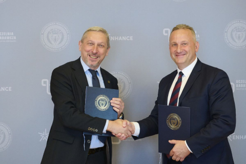Dwóch mężczyzn w garniturach pozuje do zdjęcia na tle ścianki z logo Politechniki Warszawskiej. Mężczyźni ściskają sobie dłonie, a w drugich rękach trzymają granatowe teczki z godłem uczelni.