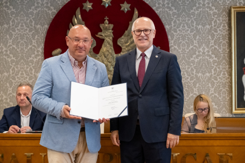 The man on the left is holding an open diploma. In the background, two seated people can be seen, along with a large WUM emblem — a golden eagle on a red background.