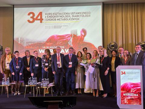 A group of several people poses on a conference stage in front of a large screen displaying the title of the “34th Continuing Education Course in Endocrinology, Diabetology and Metabolic Diseases.” Some participants are wearing conference badges on lanyards, and two people in the center of the group are holding bouquets of flowers. In front of the stage are chairs and a table with water bottles, while a lectern featuring the event branding stands on the right side.
