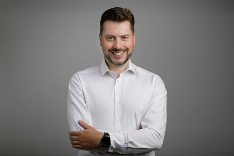 Młody mężczyzna w białej koszuli pozuje do zdjęcia portretowego. Uśmiecha się.