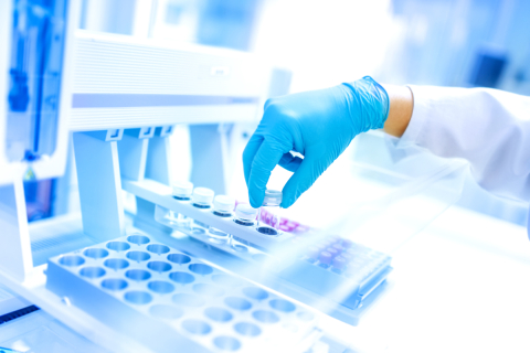 Ręka w rękawiczce, ubrana w fartuch laboratoryjny, delikatnie umieszcza fiolkę z próbką w stojaku w jasnym, czystym pomieszczeniu laboratoryjnym. Na pierwszym planie i w tle widać sprzęt laboratoryjny oraz tacki na fiolki.