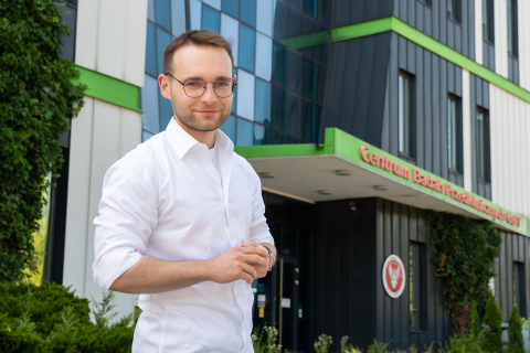 Młody mężczyzna, Karol Momot, pozuje do zdjęcia. W tle widać budynek z czarno-białą fasadą z elementami zieleni, czerwonym napisem: Centrum Badań Przedklinicznych CePT oraz godłem WUM.