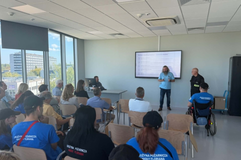 Uczestnicy spotkania siedzą na krzesłach w sali szkoleniowej i słuchają prezentacji prowadzonej przez dwie osoby stojące przed ekranem. Wśród słuchaczy znajdują się osoby ubrane w niebieskie koszulki z napisem „Drużyna Pożarnicza Anioły Floriana”, a jedna z nich siedzi na wózku. Duże okna sali wychodzą na zabudowę kampusu.