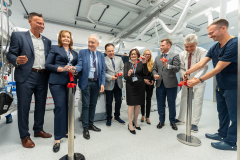 Eight people cut a red ribbon during the opening of a modern medical procedure room equipped with specialized medical equipment.