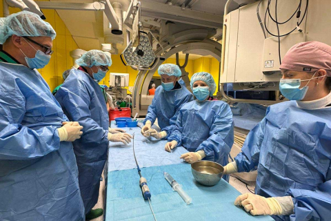 Grupa medyków w czasie zabiegu na sali operacyjnej. Wszyscy ubrani są w niebieskie fartuchy i czepki ochronne. Przed nimi na stole widać sprzęt medyczny. Za nimi widać aparaturę medyczną.
