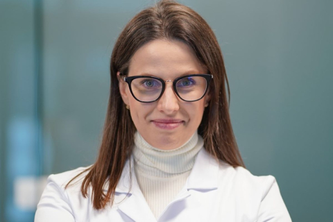 Portret osoby w białym fartuchu i ciemnych okularach, z długimi włosami, na tle rozmytego wnętrza. Osoba patrzy w obiektyw.