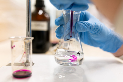 Pracownik laboratorium w niebieskich rękawiczkach dodaje fioletowy roztwór pipetą do kolby stożkowej z przezroczystą cieczą. Obok stoi zlewka z niewielką ilością różowego płynu. Widoczne są naczynia laboratoryjne oraz butelka z odczynnikiem w tle.