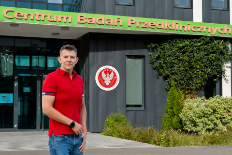 Mężczyzna, Marek Konop, pozuje do zdjęcia przed budynkiem z szarą fasadą i napisem Centrum Badań Przedklinicznych. Na budynki widać też godło WUM.