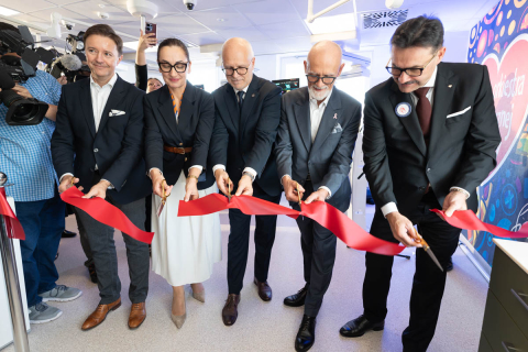 Five people jointly cut a red ribbon during the ceremonial opening of a room. Each person is holding scissors pointed towards the ribbon. On the left, a television camera and people documenting the event can be seen. In the background, there are interior furnishings and part of a colourful wall graphic.