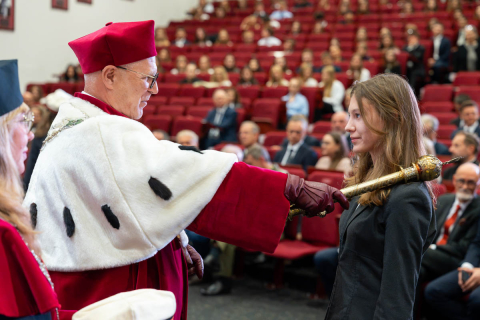 The Rector, wearing an academic gown, touches a female student’s shoulder with the ceremonial mace.