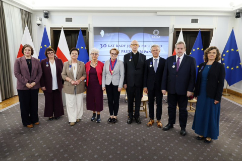 Osiem osób stoi w rzędzie podczas uroczystości w sali reprezentacyjnej. Każda osoba ma przypięte odznaczenie. W tle znajdują się flagi Polski i Unii Europejskiej oraz ekran z napisem dotyczącym 30-lecia Rady Języka Polskiego przy Prezydium PAN.