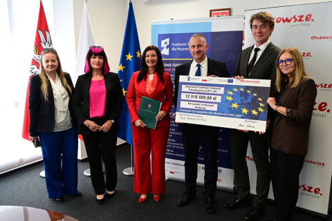 Sześć osób stoi podczas oficjalnego wydarzenia. Dwie osoby trzymają dużą planszę z informacją o dofinansowaniu projektu. W tle widoczne są flagi Polski i Unii Europejskiej oraz materiały promujące Fundusze Europejskie dla Mazowsza.