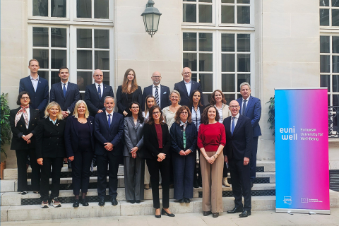 Grupa 21 osób pozuje do wspólnego zdjęcia na schodach dziedzińca. Po prawej stronie stoi baner „euniwell – European University for Well-Being”. W tle widoczne są wysokie okna i jasna elewacja budynku. Uczestnicy ustawieni są w kilku rzędach i patrzą w kierunku aparatu.
