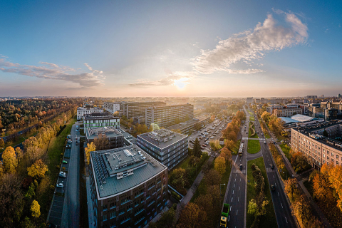 Panorama kampusu z lotu ptaka. W centrum znajdują się nowoczesne budynki biurowo-dydaktyczne, a po prawej biegnie szeroka wielopasmowa ulica z torowiskiem tramwajowym i samochodami. Po lewej stronie widoczny jest pas zieleni z drzewami. W tle rozciąga się zabudowa miasta. Nad horyzontem znajduje się nisko położone słońce i częściowo zachmurzone niebo.