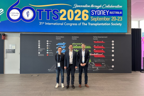 Trzy osoby pozują do zdjęcia w centrum kongresowym pod dużym ekranem informacyjnym konferencji TTS 2026 w Sydney. W tle znajduje się plan obiektu oraz oznaczenia poszczególnych stref centrum konferencyjnego