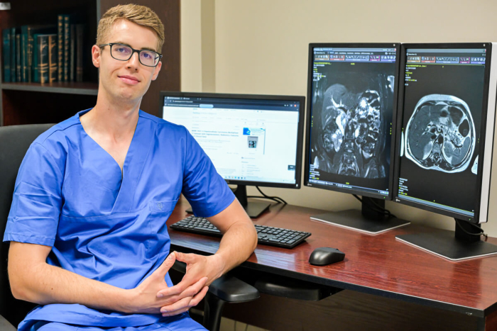 Młody mężczyzna w okularach, ubrany w niebieski fartuch lekarski. Siedzi przy stole. Na stole są trzy ekrany komputerowe. Na dwóch wyświetlają się obrazy radiologczne.