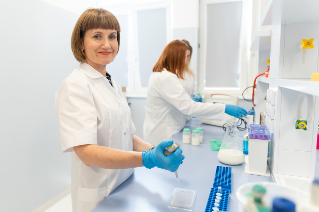 Osoba w fartuchu laboratoryjnym i rękawiczkach pracuje przy stanowisku badawczym, korzystając z automatycznej pipety. W tle druga osoba wykonuje czynności laboratoryjne przy blacie roboczym.