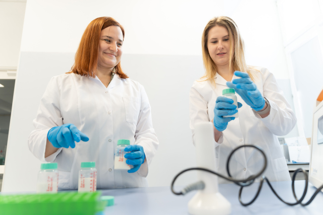 Dwie osoby w fartuchach laboratoryjnych i rękawiczkach pracują przy stole laboratoryjnym. Trzymają pojemniki z próbkami i przygotowują materiał do badań.