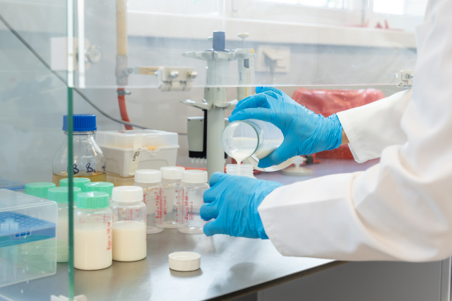 Zbliżenie na dłonie w rękawiczkach przelewające płyn do pojemnika. Obok ustawione są pojemniki z próbkami oraz wyposażenie laboratoryjne.